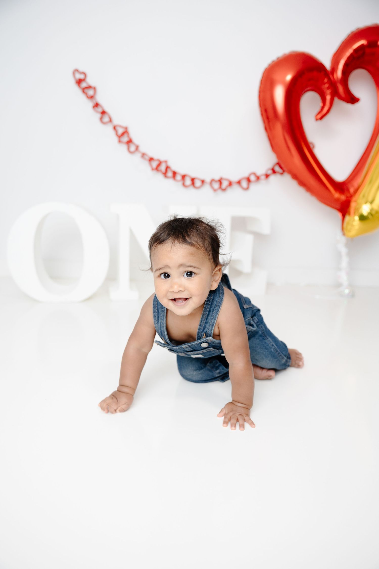 Baby girl in denim overalls crawling on white floor at first birthday party with ONE letters and heart balloons.