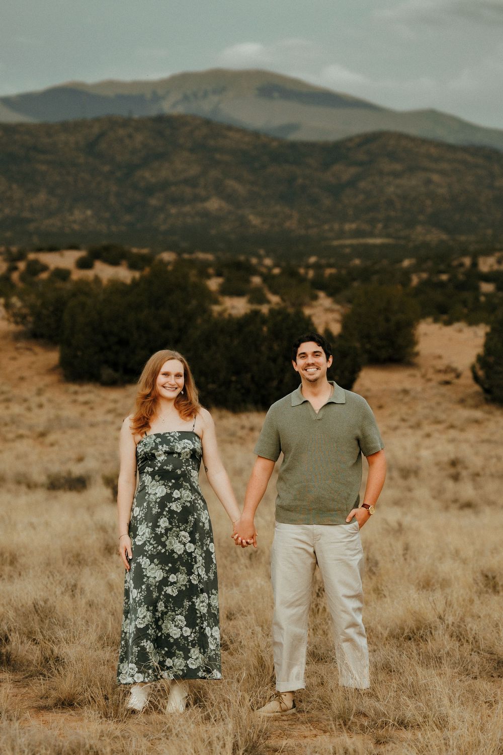 Couple holding hands in a field with mountain backdrop, woman in floral dress and man in green polo shirt.