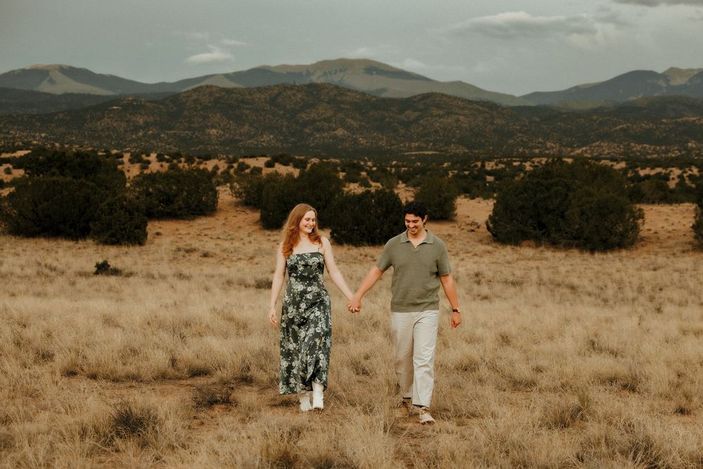 Couple holding hands walking through golden meadow with mountain backdrop during outdoor engagement photo session.