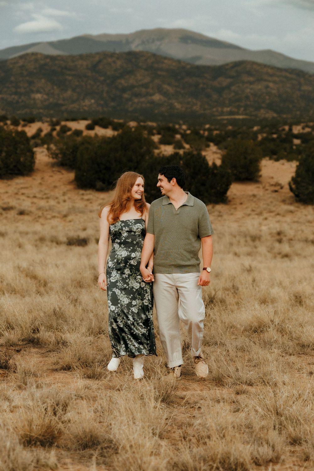 A couple walks hand-in-hand through a golden meadow with mountain views, she in a floral dress, he in a green polo.