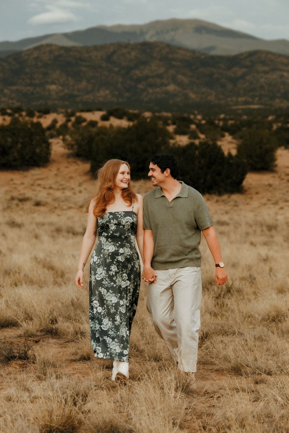 Couple holding hands walking through a golden field with mountain views in the background.