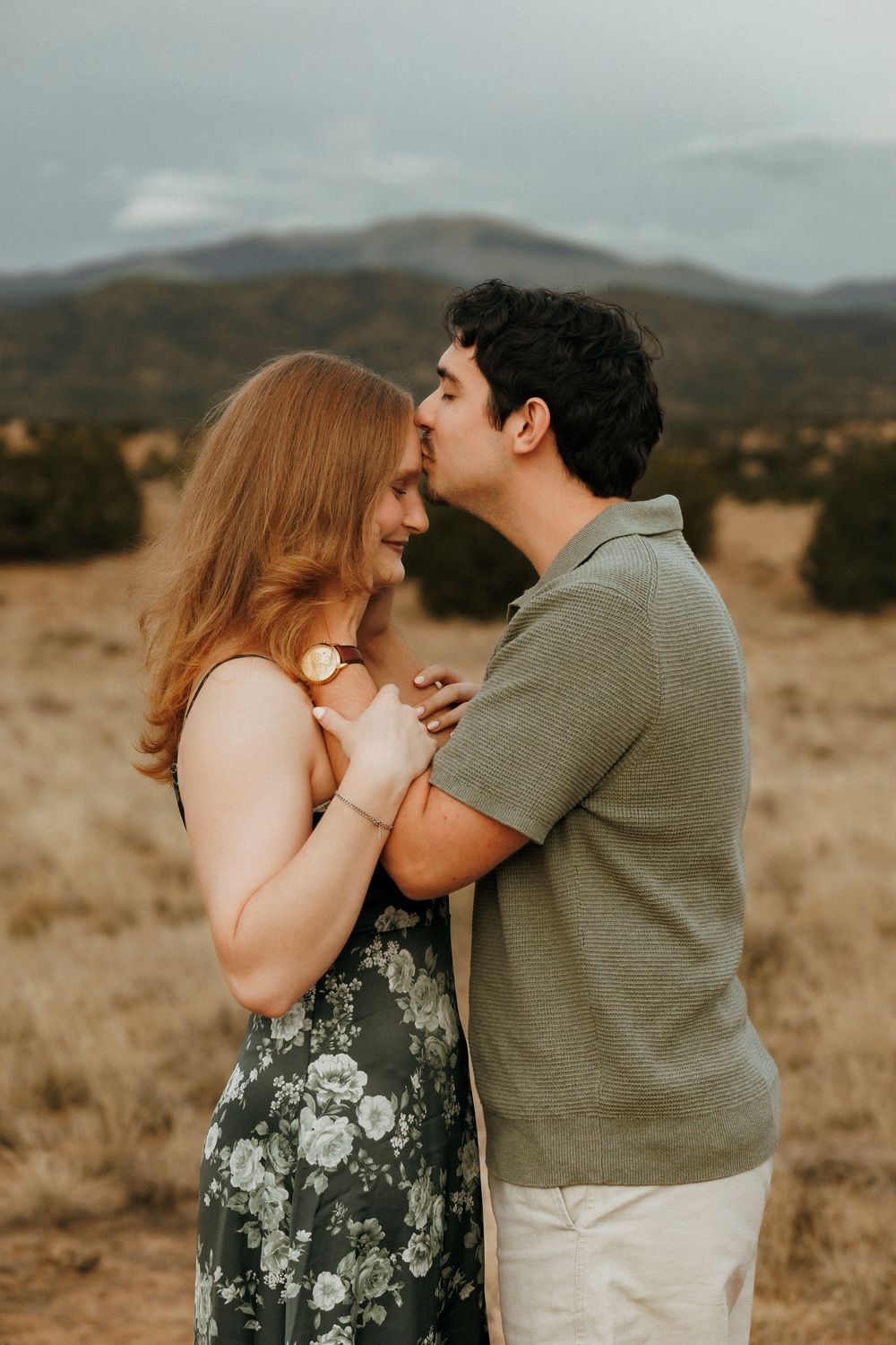 A couple embraces in a golden field with mountain backdrop, man kissing womans forehead in romantic outdoor portrait.