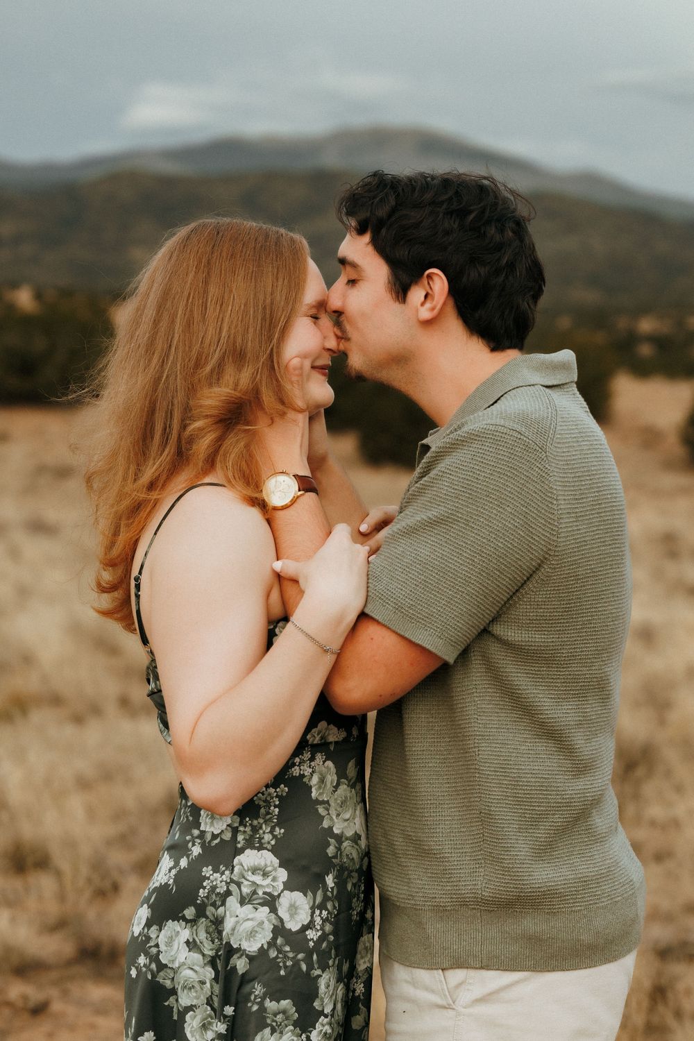 A couple shares a romantic moment in a golden field with mountain backdrop, woman in floral dress smiling.