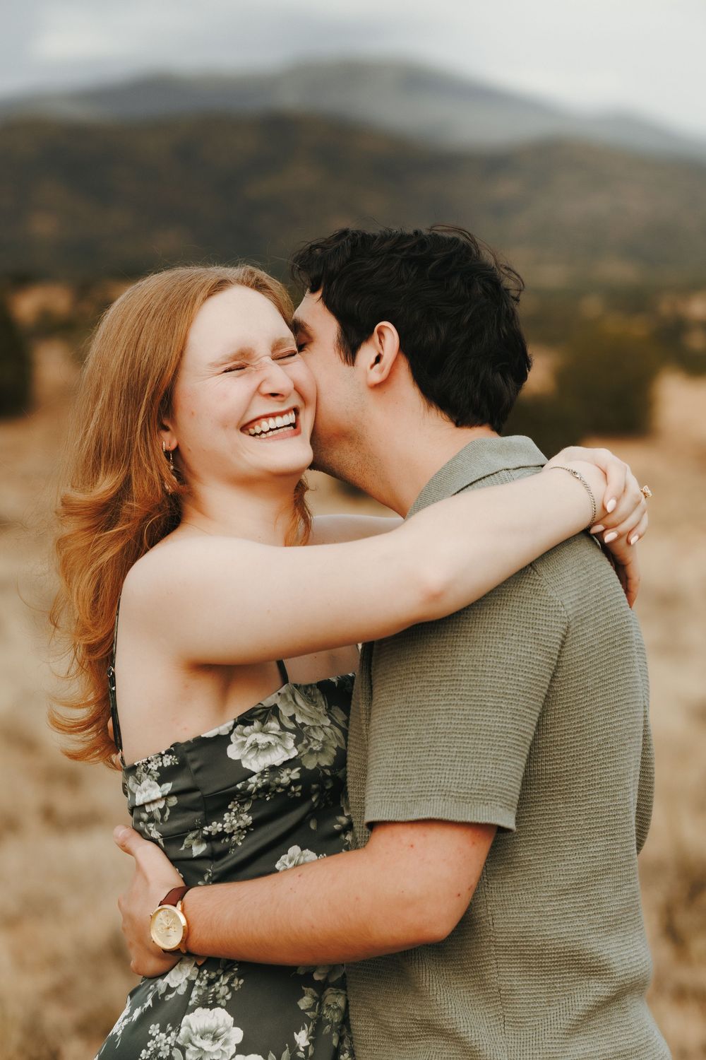 A joyful couple embraces outdoors, man kissing woman's cheek as she laughs, mountains in the background.