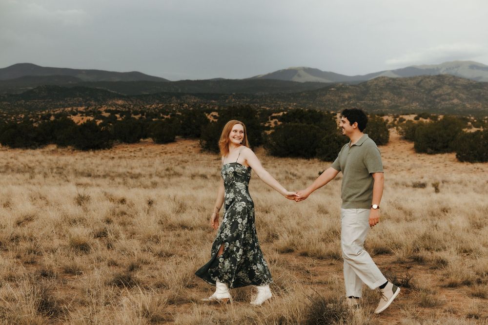 Couple holding hands walking through golden meadow with mountain backdrop under cloudy sky.