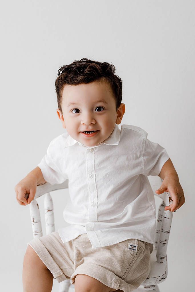 Smiling toddler boy in white linen shirt and beige shorts sitting on a white chair against a light background.