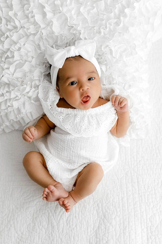 Adorable baby girl in white romper and bow headband lying on white textured blanket.
