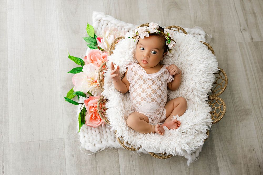 Newborn baby in white lace outfit lying in wicker basket with fur lining and pink floral wreath on head.