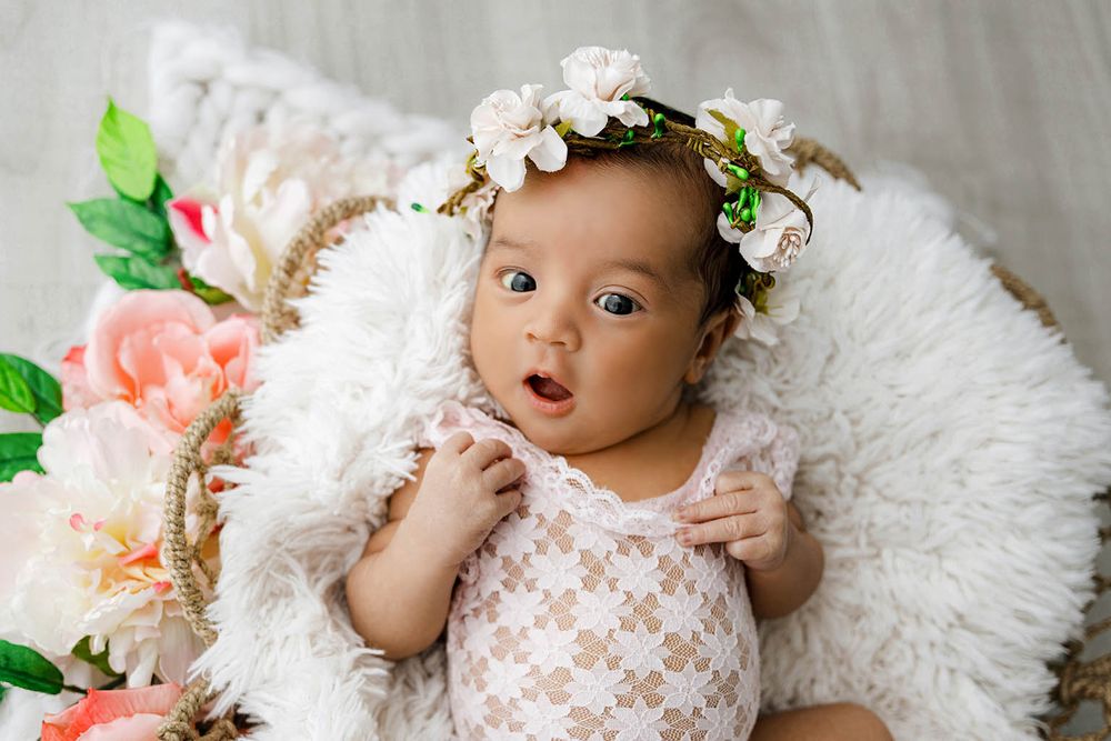 Sweet baby girl in a lace romper and floral crown lying on a fluffy white fur blanket.