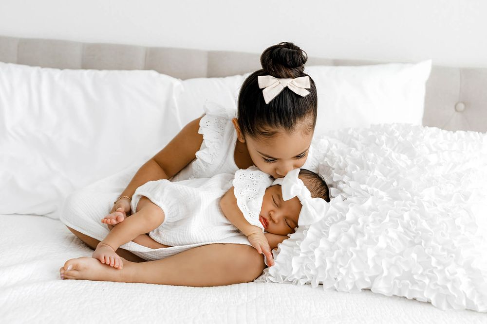 Young girl in white dress kisses newborn sibling wrapped in white on a bed.