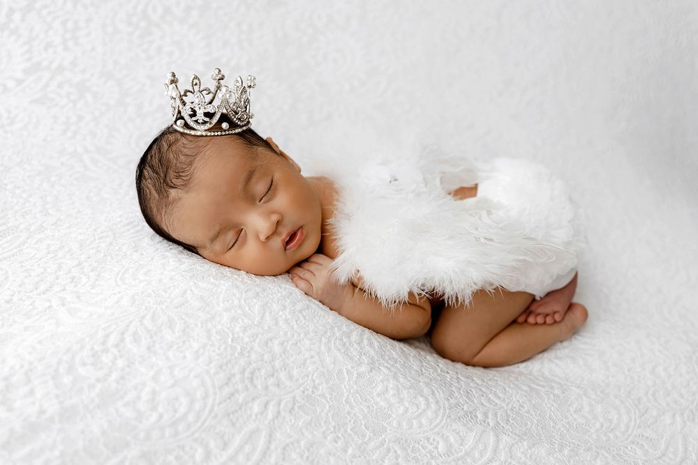 Sleeping newborn baby wearing a tiny silver crown and white feathered wings on a white blanket.