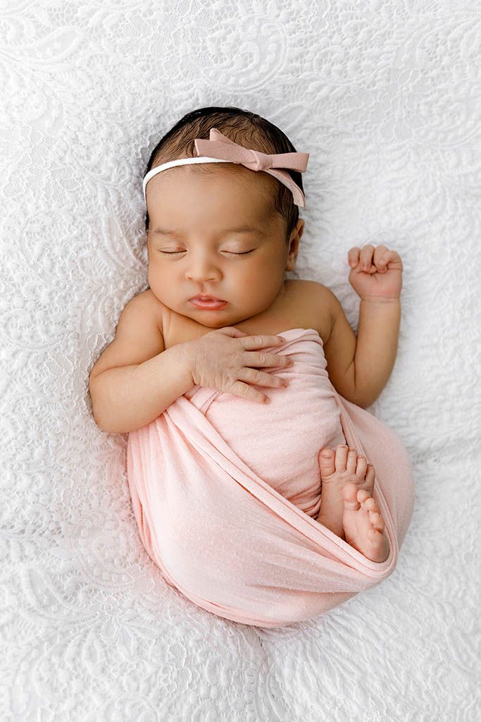 Peaceful newborn baby girl swaddled in pink wrap with bow headband sleeping on white textured blanket.