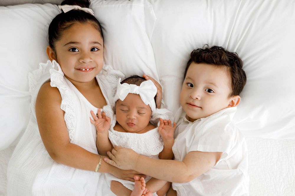 Three siblings pose on white bedding, two older children holding a sleeping newborn baby dressed in white.