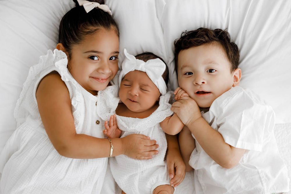 Two siblings and a newborn baby in white outfits lying together on a white bed.