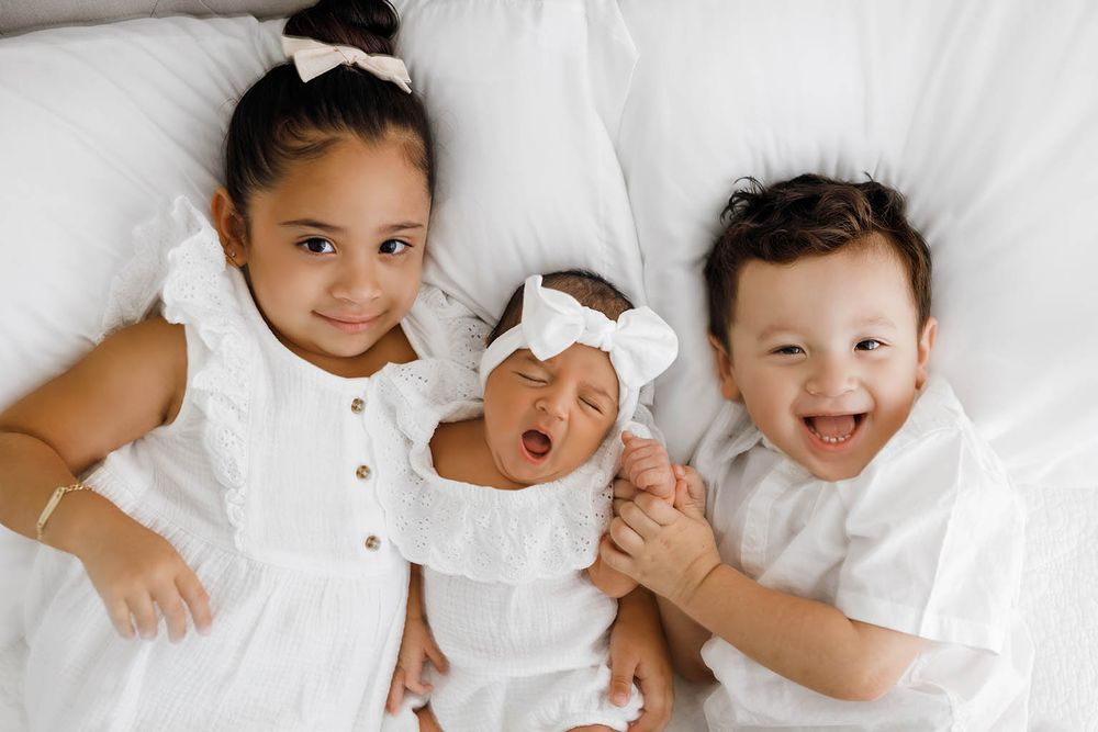 Three young siblings in white outfits lying on a white bed, with a newborn baby in the middle.