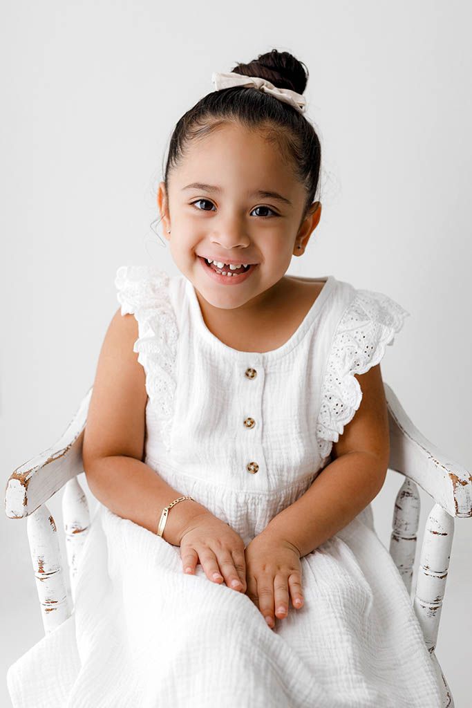 Smiling toddler girl in white dress sits on vintage chair with bow headband.