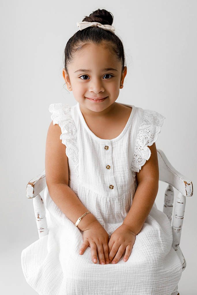 Young girl in white ruffled dress with gold buttons sits smiling on a white wooden chair.