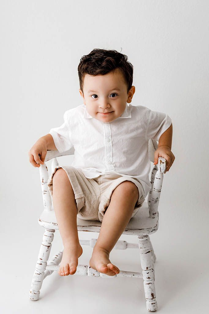Cute toddler boy in white shirt and tan shorts sits barefoot on a white wooden chair.