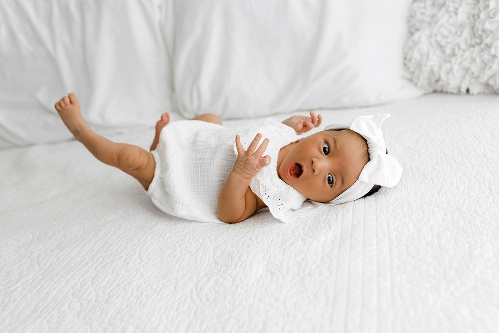 Adorable baby in white outfit and bonnet lying on white bed with mouth open in surprise.