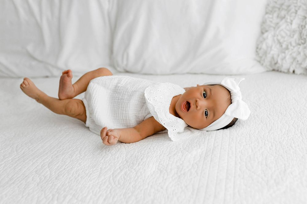 Adorable baby girl in white onesie and headband lying on a white bed, looking at the camera.