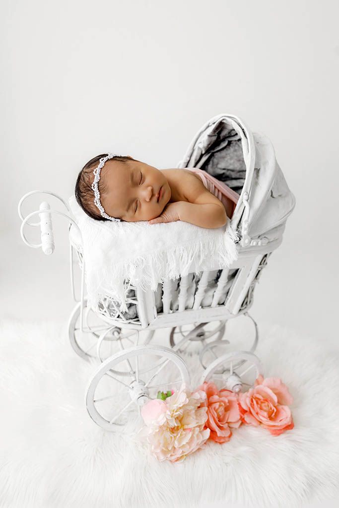 Newborn baby sleeping peacefully in a vintage white wicker pram with pink roses nearby.