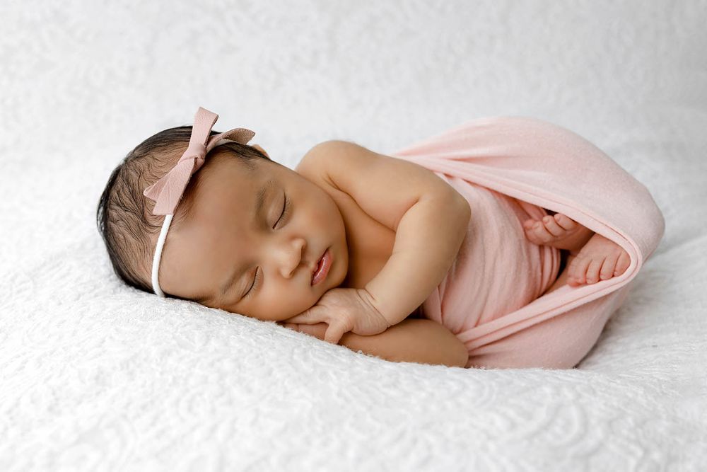 Newborn baby girl sleeping peacefully wrapped in pink swaddle with bow headband on white textured blanket.