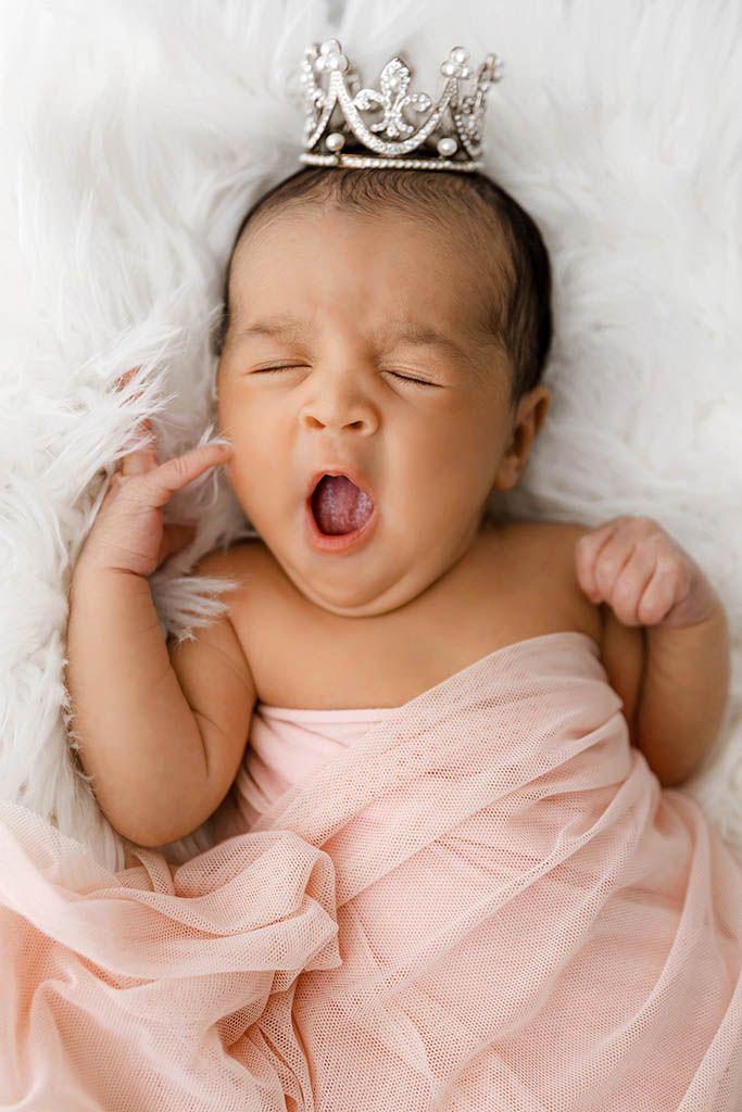 Newborn baby girl yawning, wrapped in pink tulle, wearing a tiny silver crown, lying on white fluffy blanket.