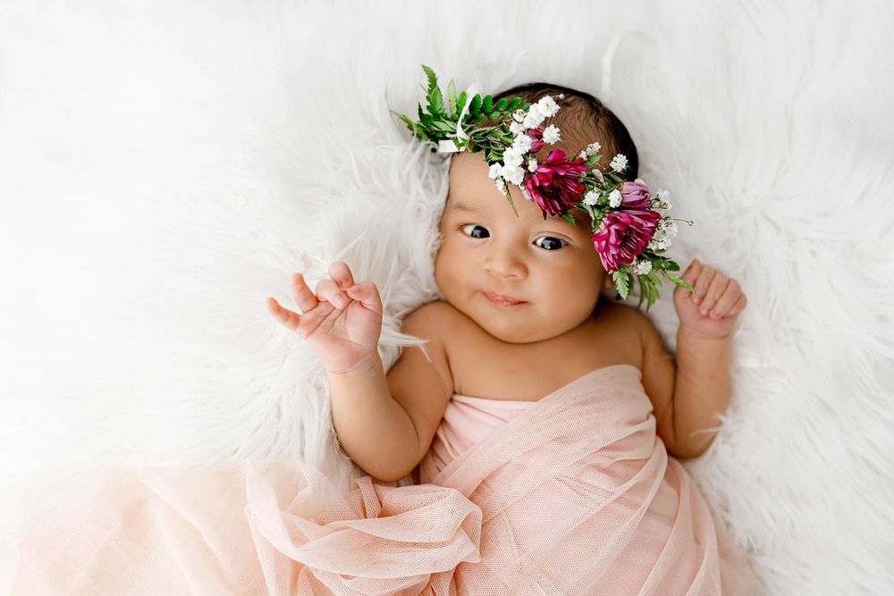 Newborn baby girl in pink wrap with floral crown lying on white fluffy fur blanket.