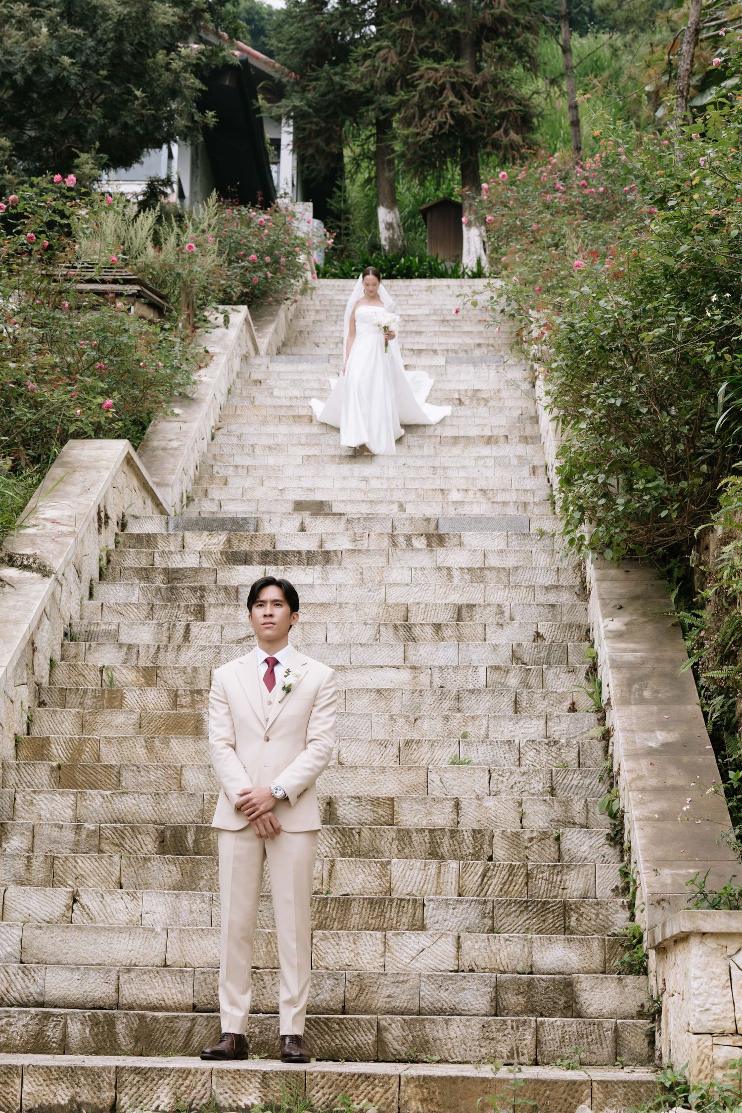 Groom in beige suit stands at bottom of stone stairs as bride in white gown descends through lush garden.
