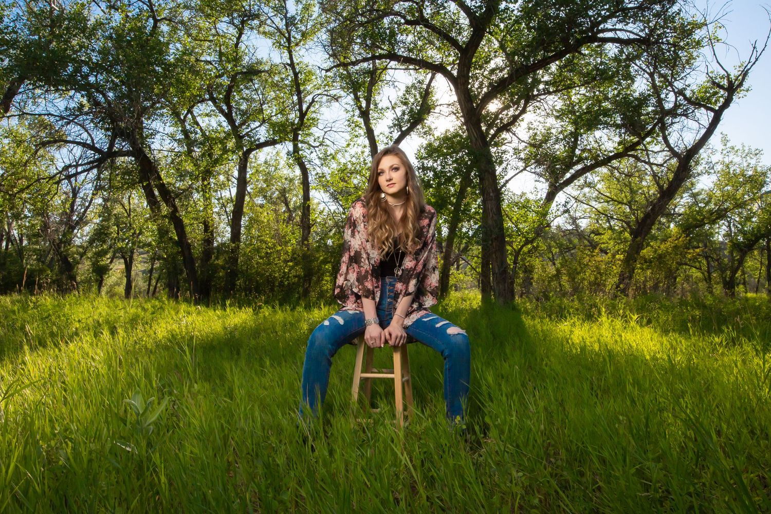 A woman sits on a wooden stool in a sunlit meadow surrounded by trees.
