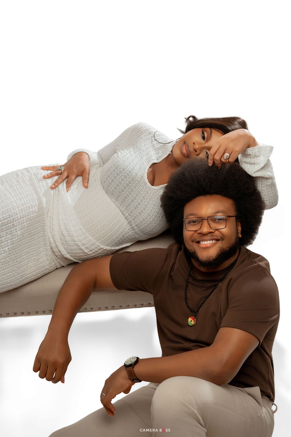 A pregnant woman in a white dress reclines on a bench while a smiling man in brown sits beside her.