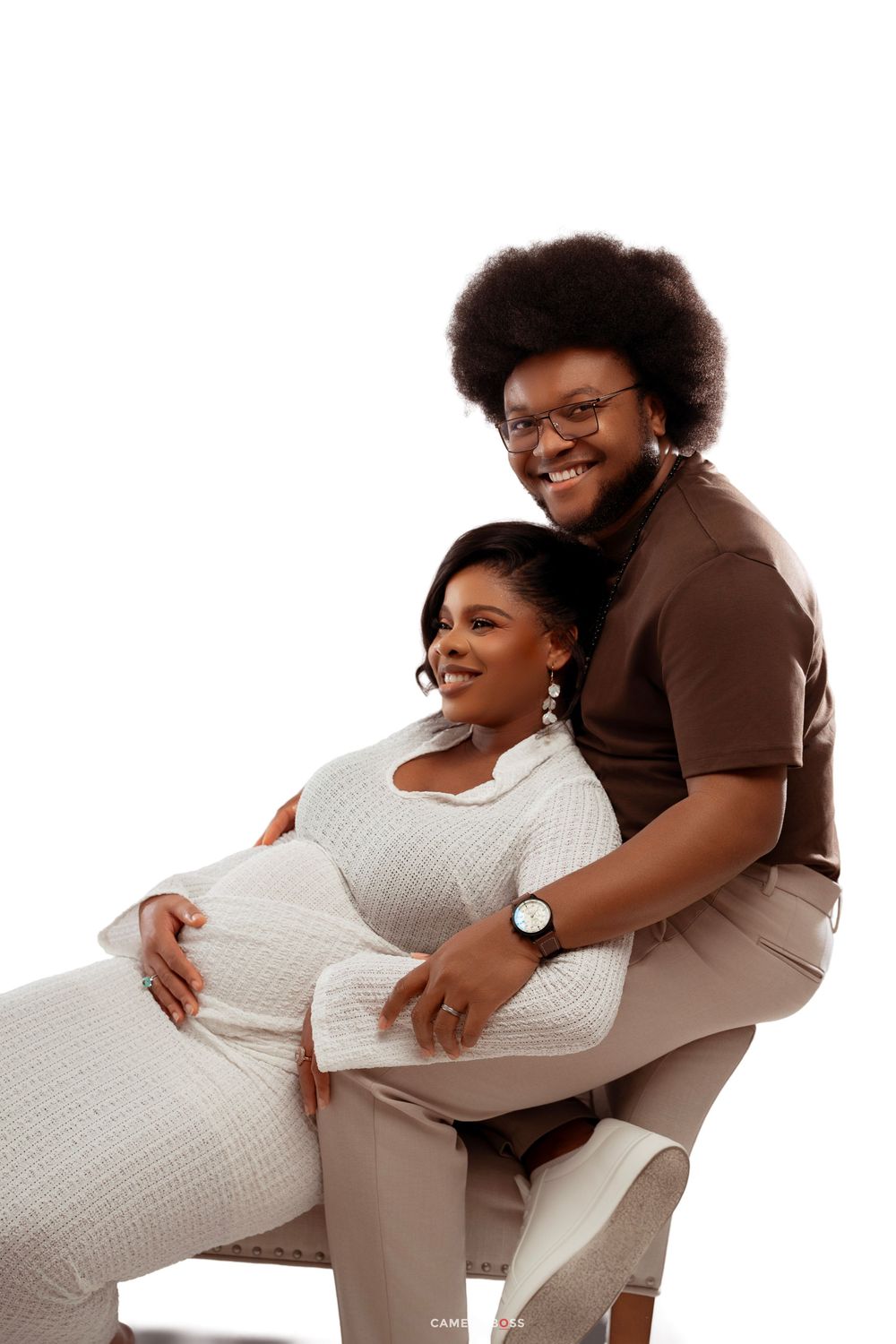 A smiling couple poses together, with a pregnant woman in white cradled by a man in a brown shirt.