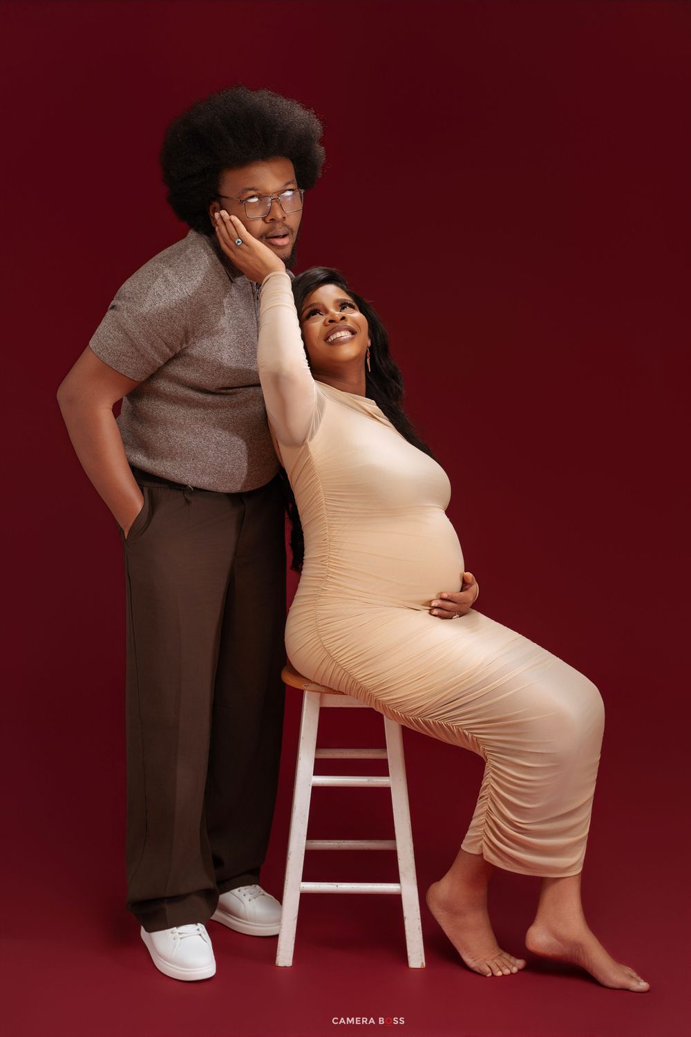Couple maternity photo with pregnant woman in nude dress seated on stool leaning on partner against red backdrop.