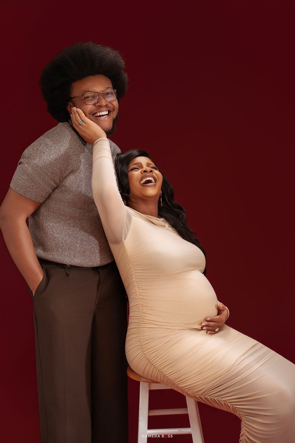 A laughing pregnant woman in a cream dress sits beside a man with an afro against a dark red background.