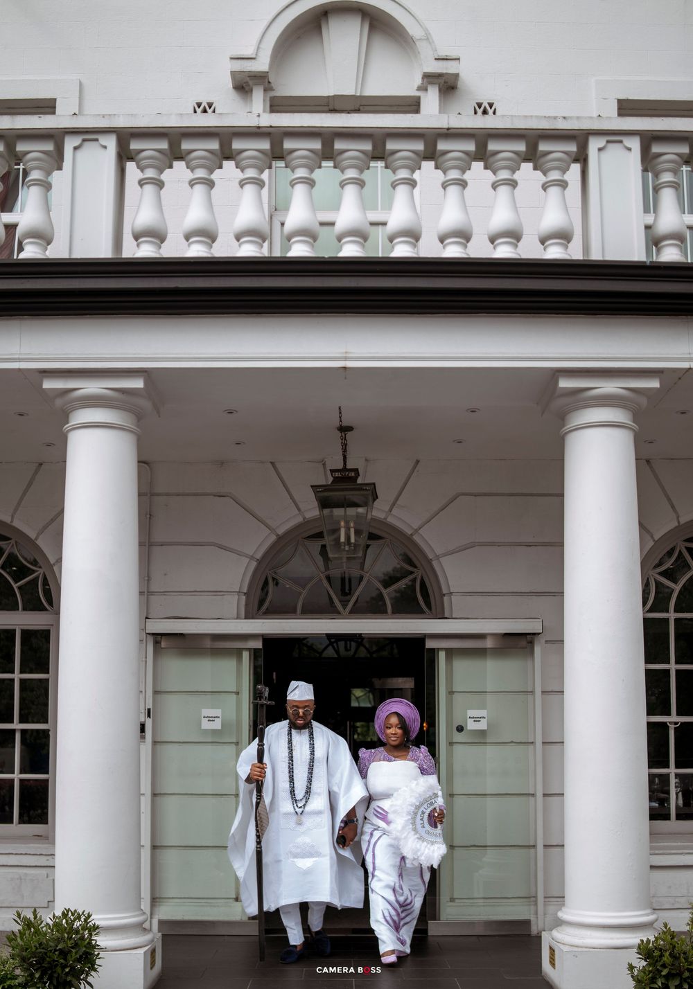 Nigerian couple in traditional Yoruba attire exit elegant white colonial building with columns during wedding ceremony.