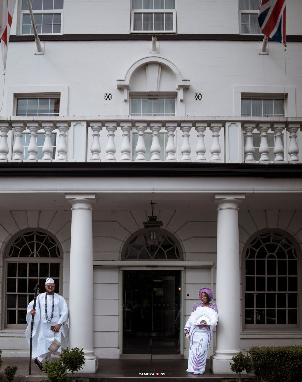 Two people in traditional white African attire stand at the entrance of an elegant white colonial building with columns.