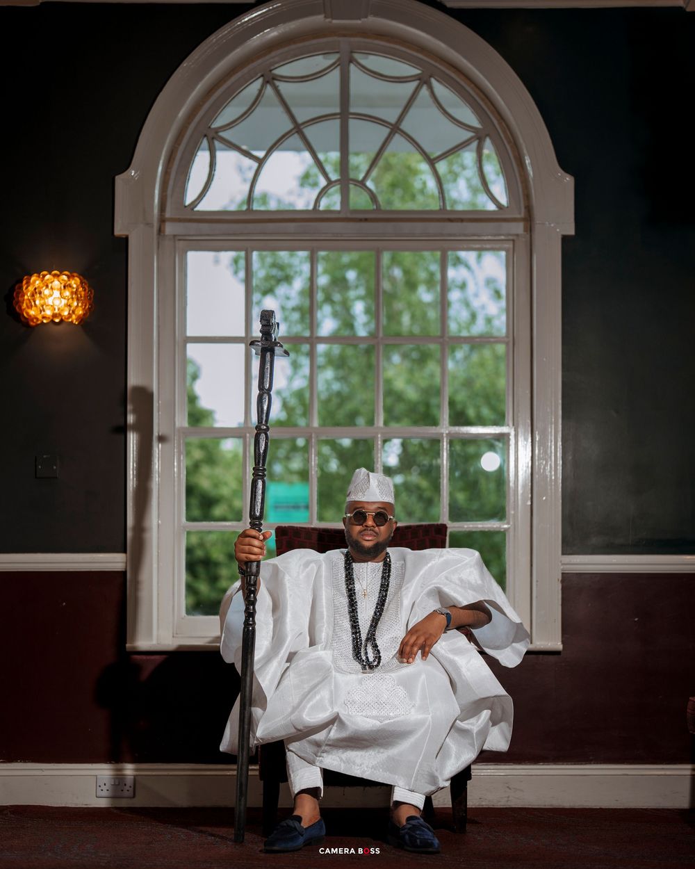 Man in white traditional Nigerian agbada attire holding a staff, seated before an arched window.