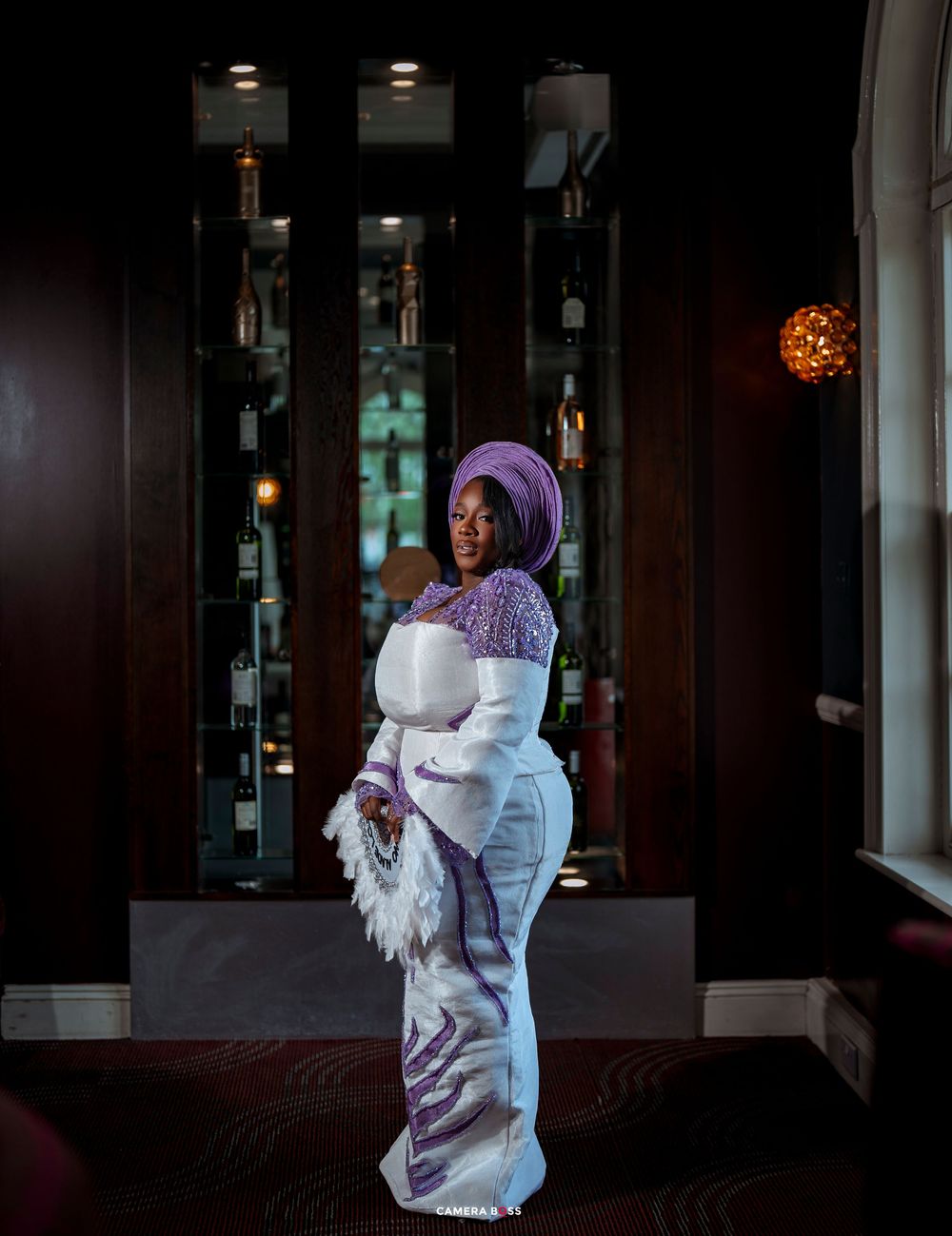 Woman in elegant white and purple traditional African attire with gele headwrap poses in a dimly lit bar.