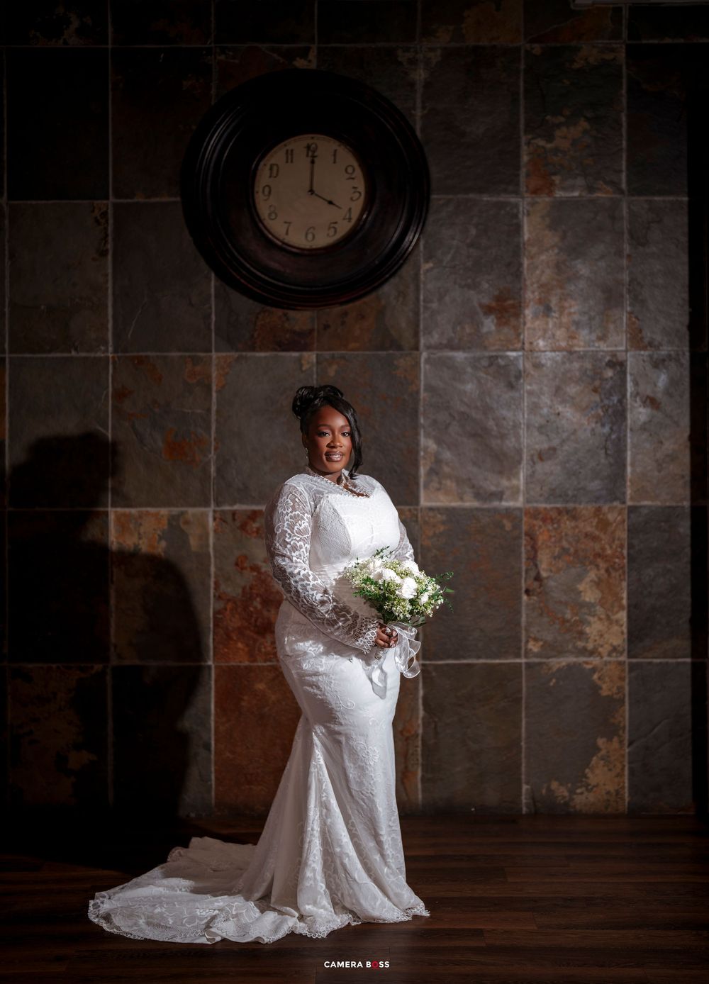 Bride in white lace gown holding bouquet stands before slate wall with clock in elegant wedding portrait.