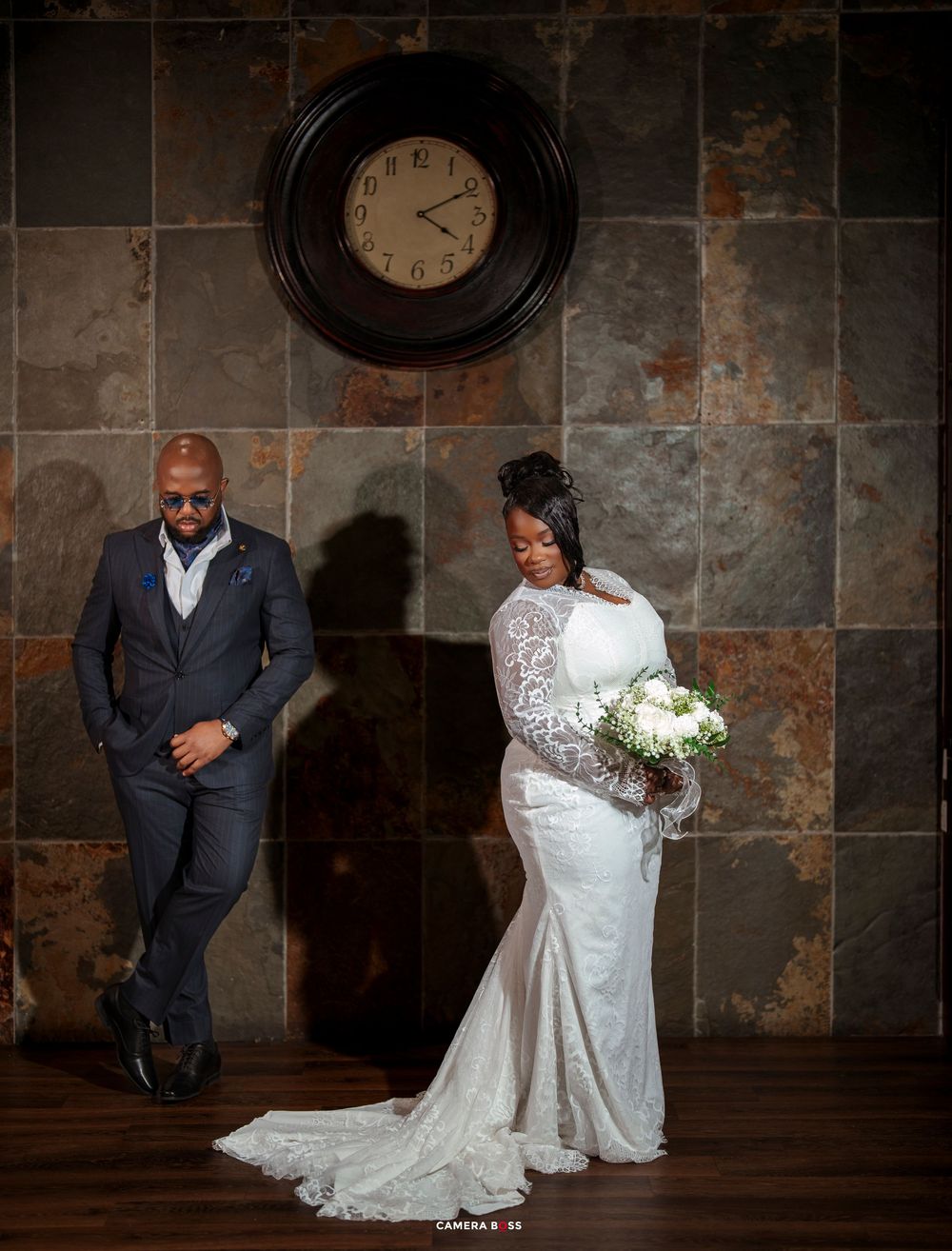 Elegant bride in lace gown holds white bouquet beside suited groom near rustic tile wall with vintage clock.