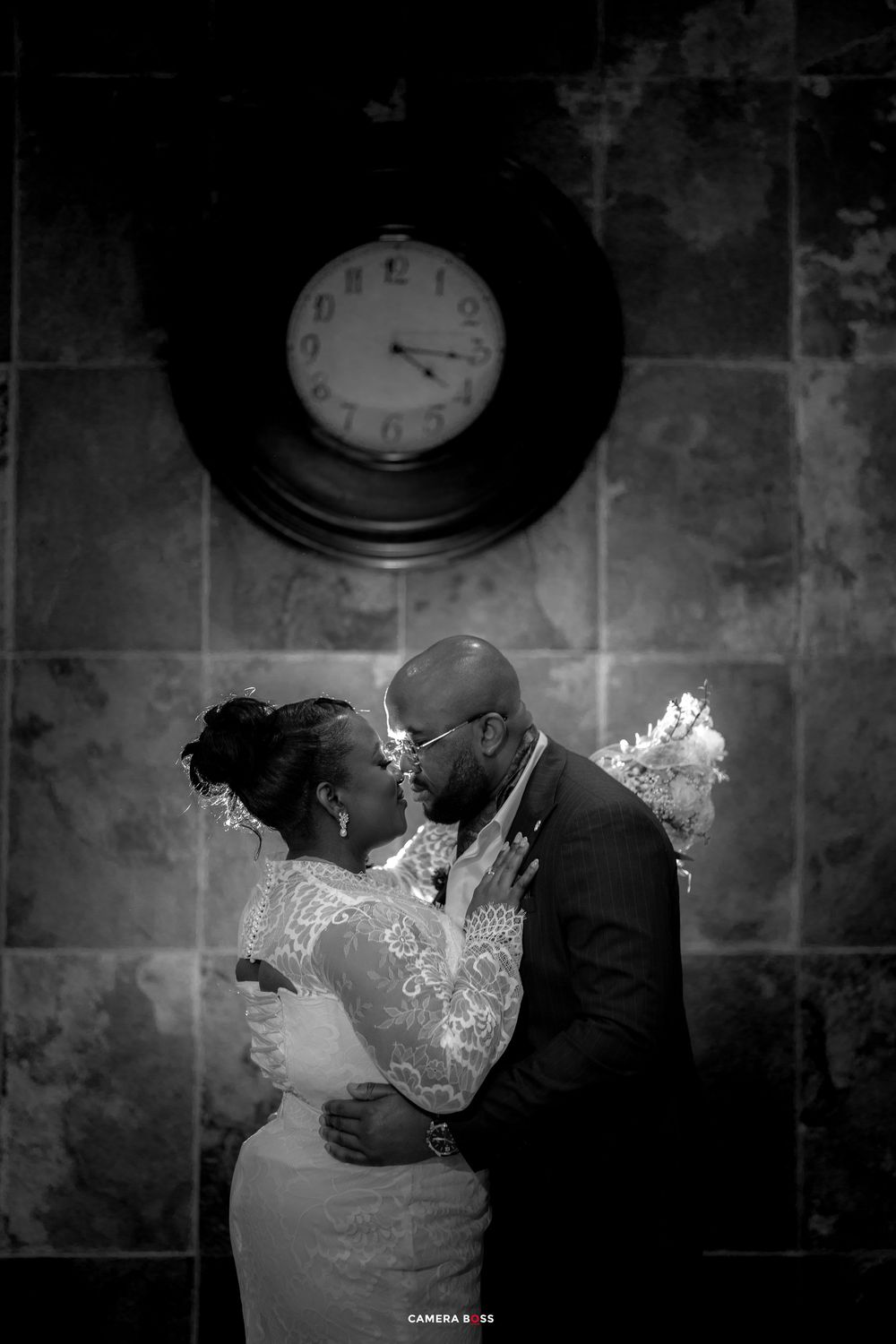 Black and white wedding photo of a couple sharing a romantic moment beneath a vintage clock on a tiled wall.