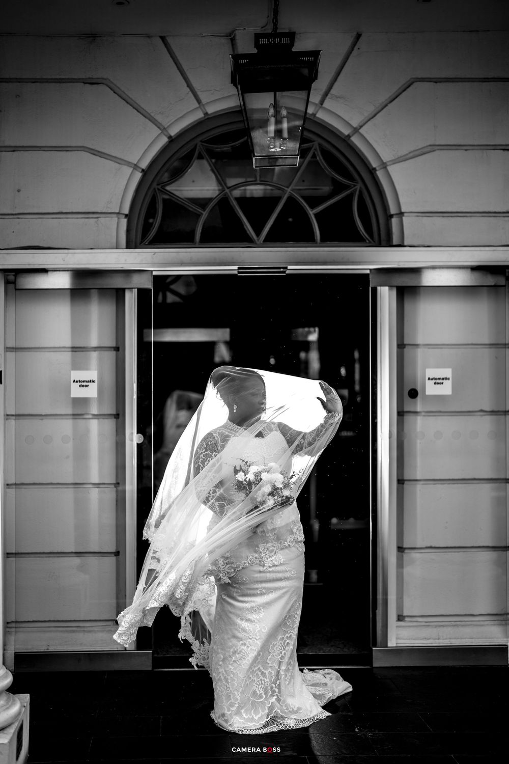Black and white photo of a bride in lace gown with veil blowing in wind, holding bouquet outside elegant building entrance.