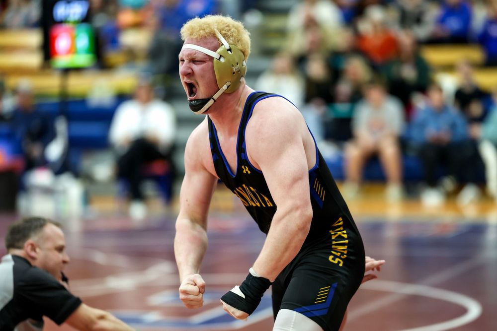 Wrestler in blue and gold singlet celebrates victory on mat, wearing headgear, pumping fist with intense expression.