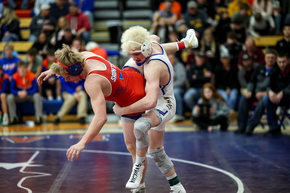 Two wrestlers compete in a high school wrestling match, one in red and one in white, grappling on the mat.