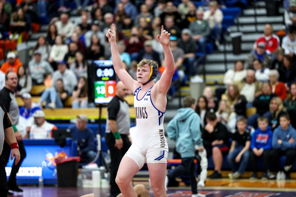 Wrestler in blue singlet celebrates victory with arms raised at a packed indoor wrestling tournament.