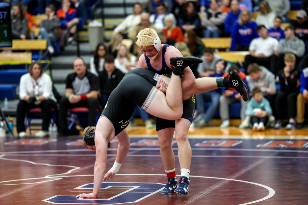 Two wrestlers compete on a blue mat during a high school wrestling match in a gymnasium filled with spectators.