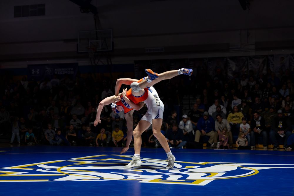 Two wrestlers compete on a blue mat during an indoor wrestling match with a crowd watching in the background.