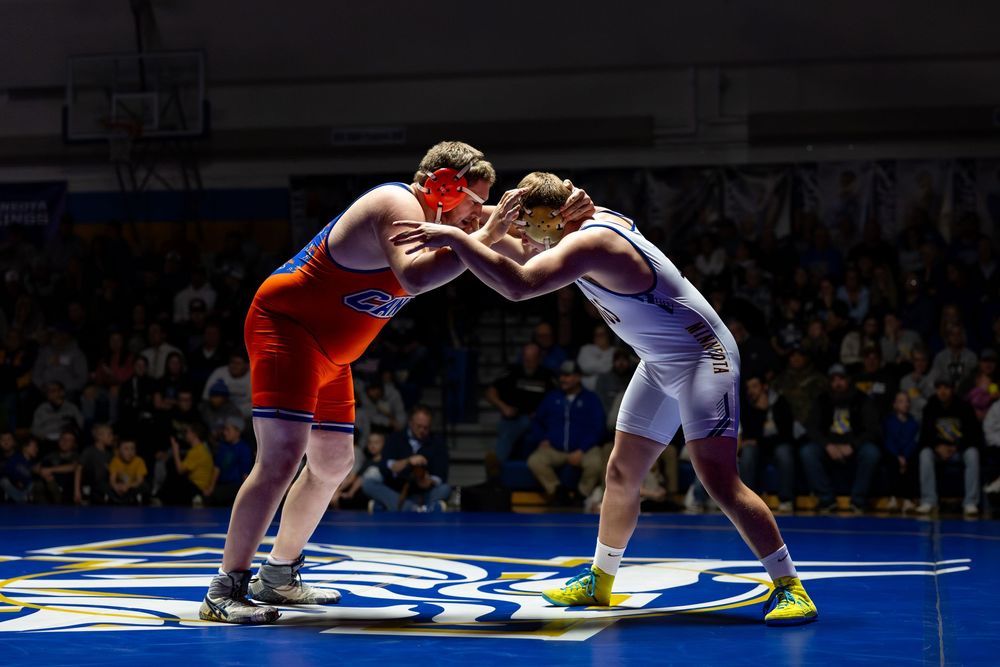 Two wrestlers compete in a college wrestling match on a blue mat under bright arena lights.