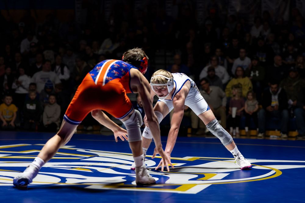 Two wrestlers compete on a blue mat in a packed arena, one in orange and the other in white uniforms.