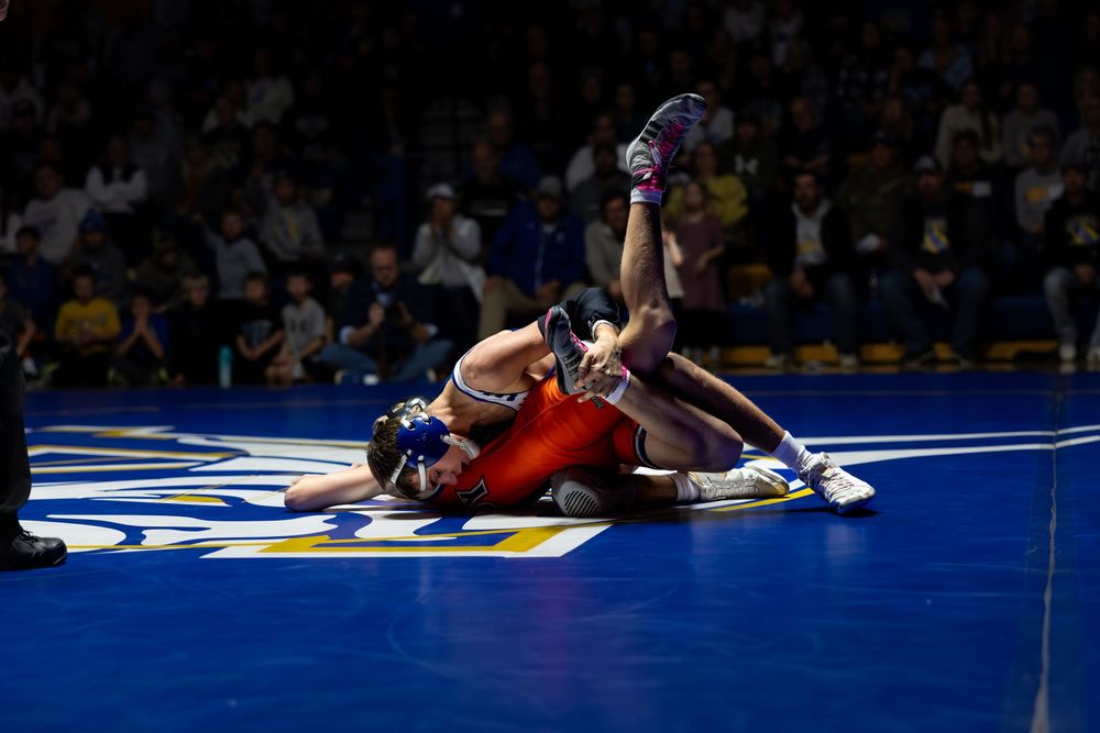 Two wrestlers compete intensely on a blue mat during a college wrestling match under bright arena lights.